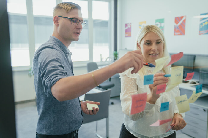 Zwei Personen arbeiten zusammen an einem Whiteboard mit bunten Haftnotizen in einem Büro.