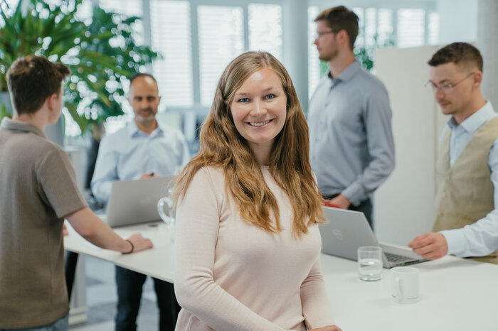 Eine Frau lächelt in die Kamera, während im Hintergrund mehrere Menschen an einem Tisch mit Laptops arbeiten.
