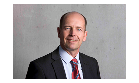 Ein Mann in einem Anzug mit Krawatte steht vor einem grauen Hintergrund und lächelt in die Kamera.