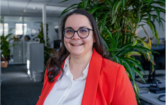 Eine Person mit Brille und rotem Blazer lächelt in einem Büro mit Pflanzen im Hintergrund.