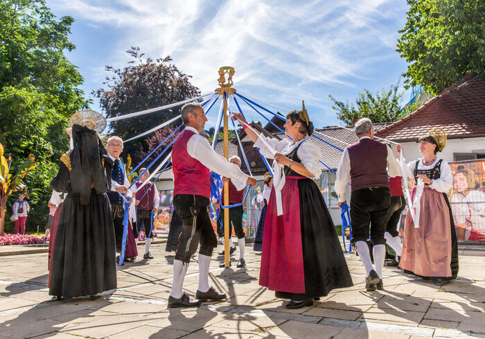 Menschen in traditioneller Tracht tanzen um einen Maibaum mit bunten Bändern.
