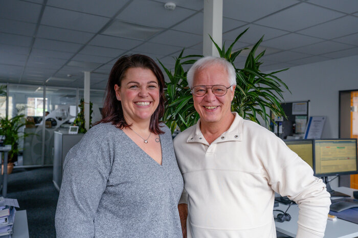 Zwei Personen lächeln in einem Büro mit Pflanzen und Computern im Hintergrund.