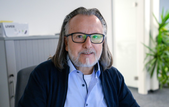 Ein Mann mit Brille und langen Haaren sitzt lächelnd in einem Büro.