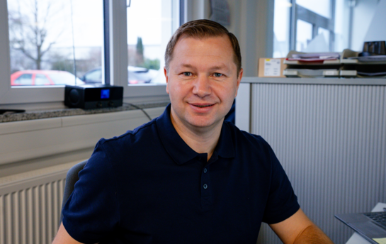 Ein Mann, ohne Bart sitzt in einem Büro an einer Fenster-Front.