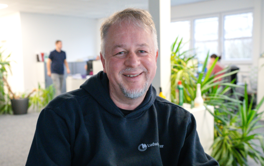 Ein älterer Mann und grauem Bart sitzt in einem Büro, umgeben von Pflanzen. | © Freundlicher Mann im Büro