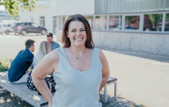 Ein Frau im weißen Top lacht in die Kamera, während man im Hintergrund Personen sieht, die sich unterhalten. 