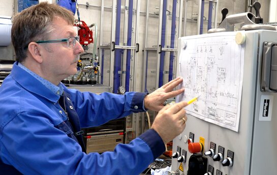 Ein Ingenieur in blauer Arbeitskleidung überprüft technische Zeichnungen an einem Schaltschrank.